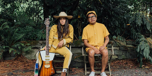 A picture with green tropical plants and trees in the background and two people dressed in yellow sit next to each other on wooden chairs. Person on left is holding a brown guitar and a yellow hat while the person on right wears a yellow beret.