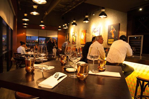 A rustic wooden table is set for McAllen drink and dining with four wine and copper glasses while other patrons enjoy their time at the nearby bar and tables. The McAllen restaurant is dimly lit with vintage posters adorning the walls.